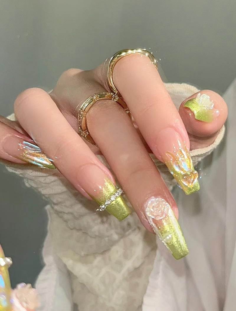 Bàn tay với móng tay được trang trí màu xanh lá và vàng, có họa tiết hoa, đeo nhẫn vàng trên ngón tay.