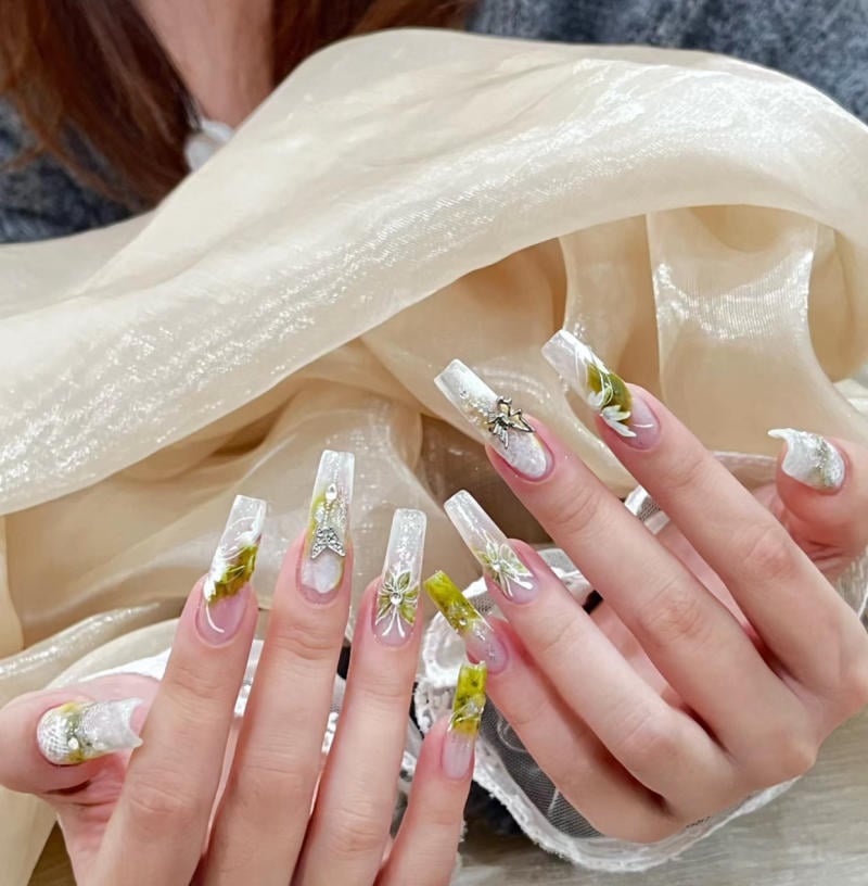 Bộ móng tay dài được trang trí với hoa văn màu xanh lá và trắng, nền vải ánh kim phía sau.