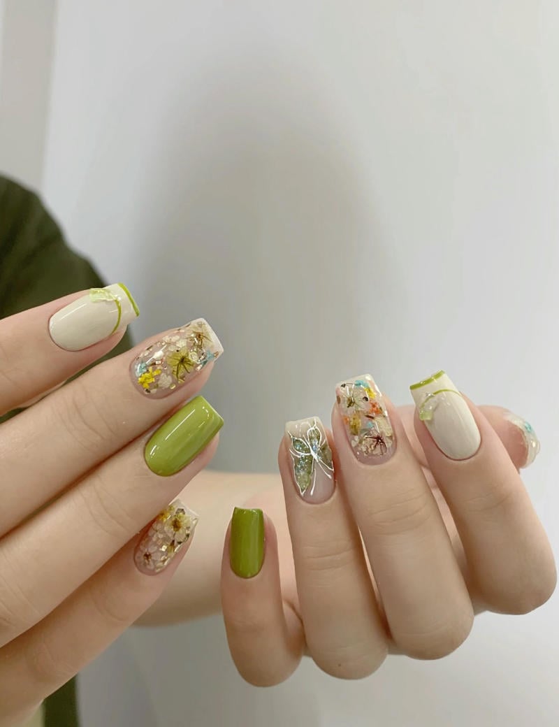 Bàn tay với móng tay trang trí hoa văn, màu xanh lá và họa tiết hoa nổi bật trên nền trắng.