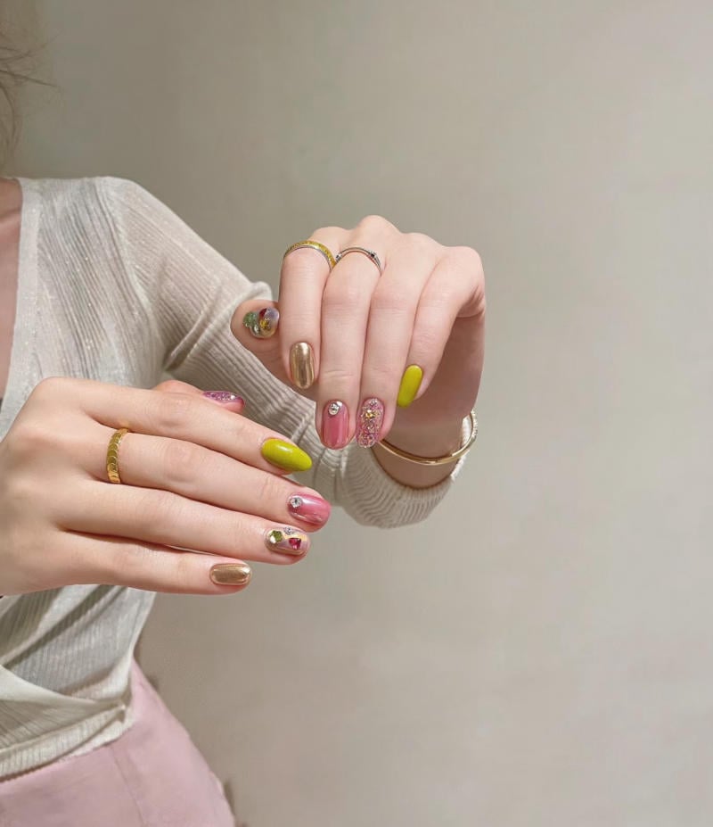 Bàn tay với móng tay được trang trí nhiều màu sắc và họa tiết, đeo nhẫn vàng, trên nền áo sáng màu.