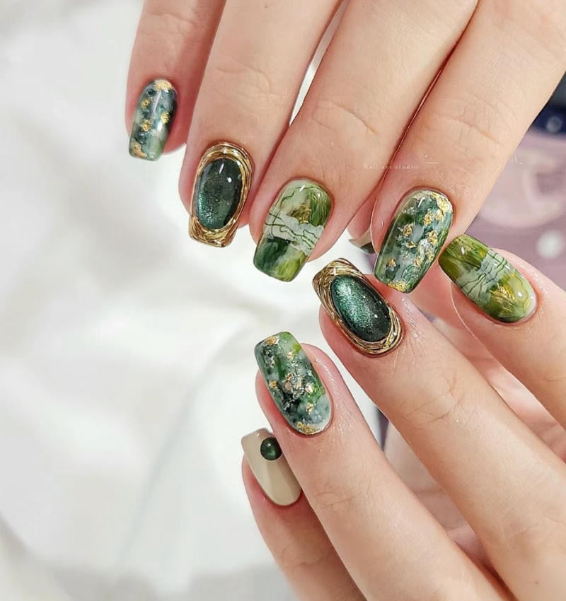 Bàn tay với móng tay được trang trí nghệ thuật, màu xanh lá đậm, họa tiết kim loại và đá quý.