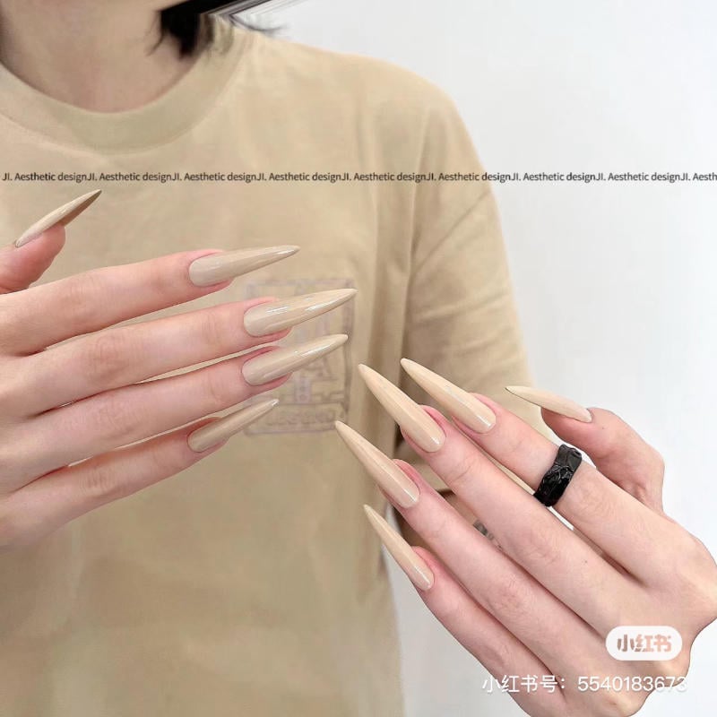 nail-mong-nhon-tron__10__3800c14e06bb45c1a346fc96bd6ec35e Bàn tay với móng tay dài, sơn màu nude bóng, một tay đeo nhẫn đen, nền áo màu be.