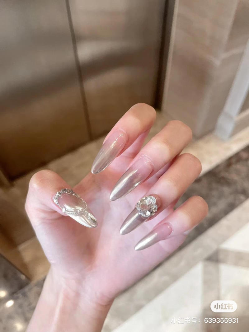 nail-mong-nhon-tron__11__db471d57cdc24ecfa82b8adf03717101 Bàn tay với móng tay dài, trang trí màu bạc và đính đá lấp lánh, nền là sàn nhà màu be.