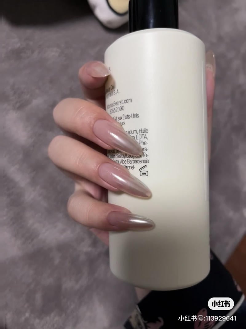 nail-mong-nhon-tron__12__fbee7b2e40cf463bb5a92de2ea067da4 Tay cầm chai mỹ phẩm màu trắng, móng tay dài và sơn bóng, nền xám mờ.