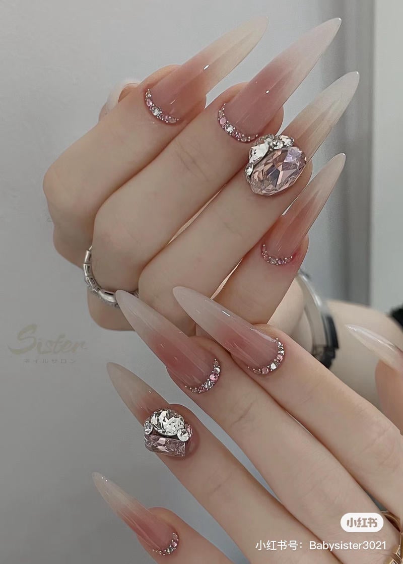 nail-mong-nhon-tron__13__12aa43f80137409c8f05c74da7a61670 Bàn tay với móng tay dài, màu nude, trang trí đá lấp lánh và nhẫn kim loại.
