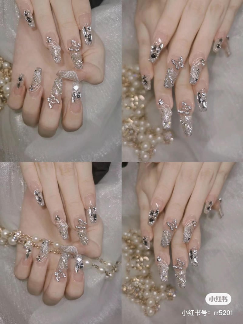 nail-mong-nhon-tron__14__efcb3674298f4286aaebd0a67e1d34ab Bàn tay với móng tay dài, được trang trí họa tiết kim loại và đá quý, đặt trên nền vải trắng và chuỗi hạt.