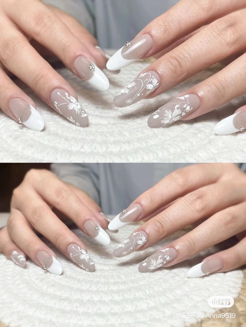 nail-mong-nhon-tron__4__5e67ae72a6924616a6db6b40bee87316 Bàn tay với móng tay dài được trang trí hoa văn trắng và đính đá trên nền màu nude và trắng.