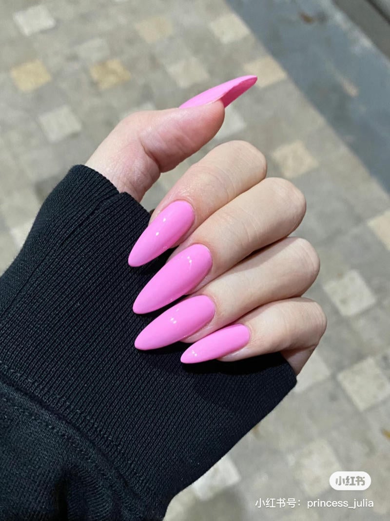 nail-mong-nhon-tron__6__70f2b33dfa514c03a876171c20bff7de Bàn tay với móng dài, sơn màu hồng sáng bóng, áo tay dài màu đen, nền gạch xám.