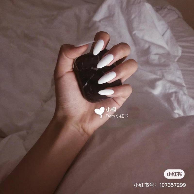 nail-mong-nhon-tron__7__e7173715703d4bbdb32791a48b9c0510 Bàn tay với móng tay sơn màu trắng cầm một vật tối màu, nền là ga giường màu trắng.