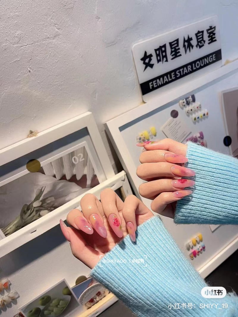 nail-mong-nhon-tron__8__2bc3f46c37354b9bb3c249ba2c819022 Hai bàn tay với móng tay được trang trí họa tiết hoa, nền có bảng hiệu và các khung hình trưng bày.