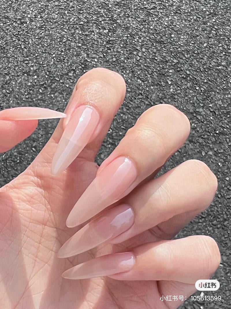 nail-mong-nhon-tron__9__1b0563d8546d4aac85e8f044eb53aad1 Bàn tay với móng tay dài, nhọn, sơn màu hồng nhạt, trên nền đá xám.