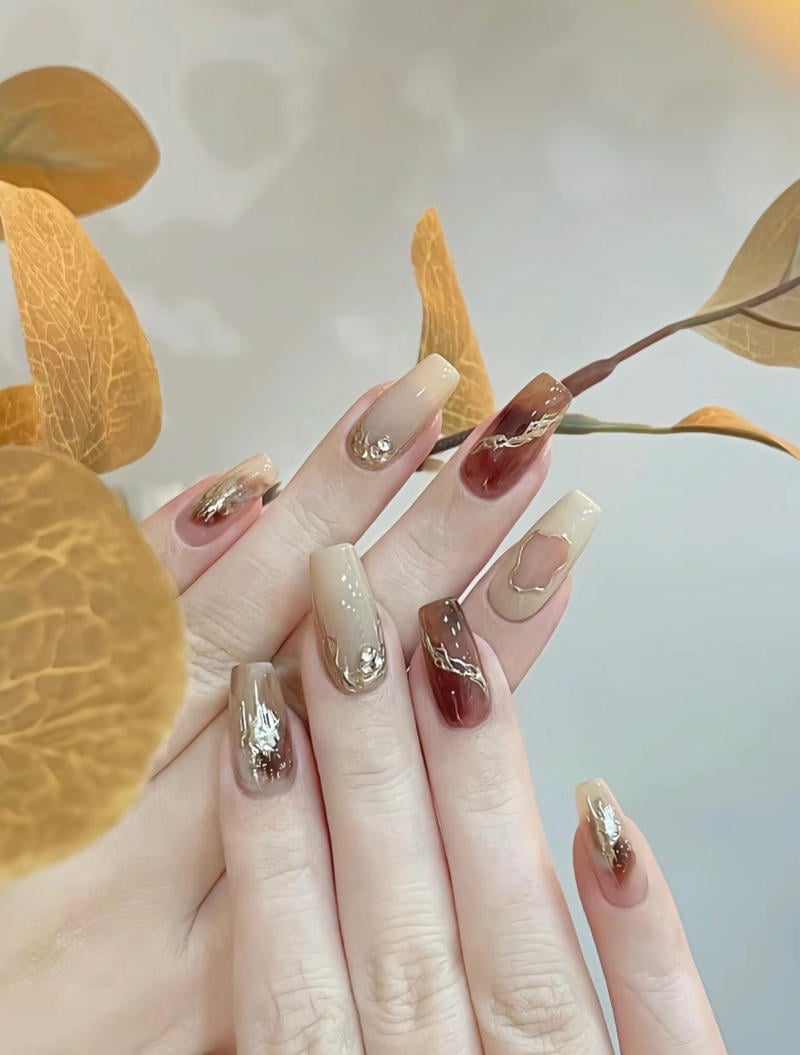 Bàn tay với móng tay được sơn màu nâu và kem, có họa tiết trang trí, bên cạnh lá cây vàng.
