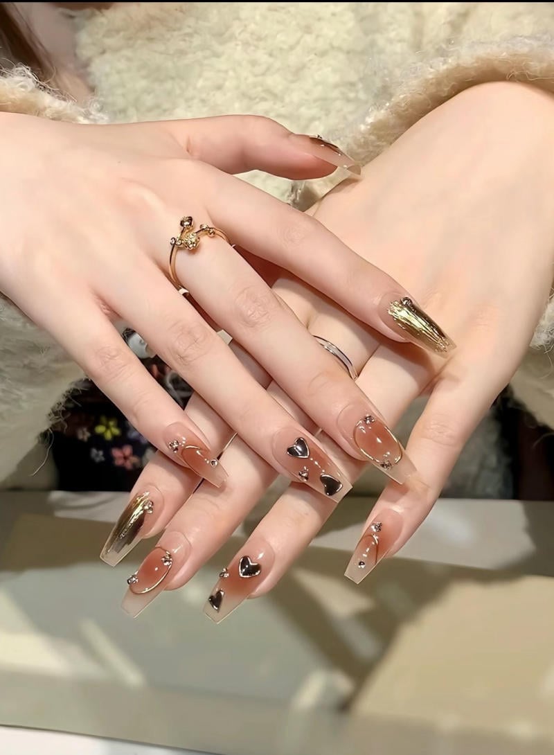 Bàn tay với móng tay đính đá và họa tiết trái tim, sơn màu nâu và vàng, đeo nhẫn trang trí.
