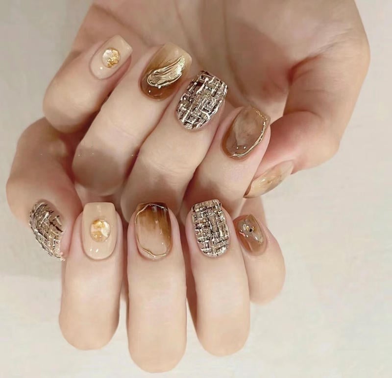 Bàn tay với móng tay được trang trí họa tiết phức tạp và màu sắc phong cách trầm, kết hợp ánh vàng và họa tiết đan.