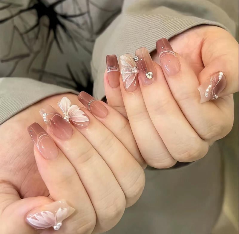 Bàn tay với móng tay được trang trí họa tiết hoa và bướm, màu sắc nhẹ nhàng và đính đá lấp lánh.