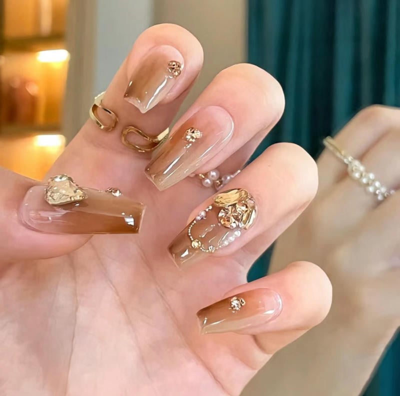 Bàn tay với móng tay sơn màu nâu, trang trí đá và hoa văn tinh tế, đeo nhẫn vàng và nhẫn ngọc trai.