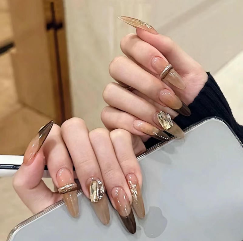 Bàn tay với móng tay dài được sơn màu nâu nhạt, trang trí đá quý, cầm điện thoại màu bạc.