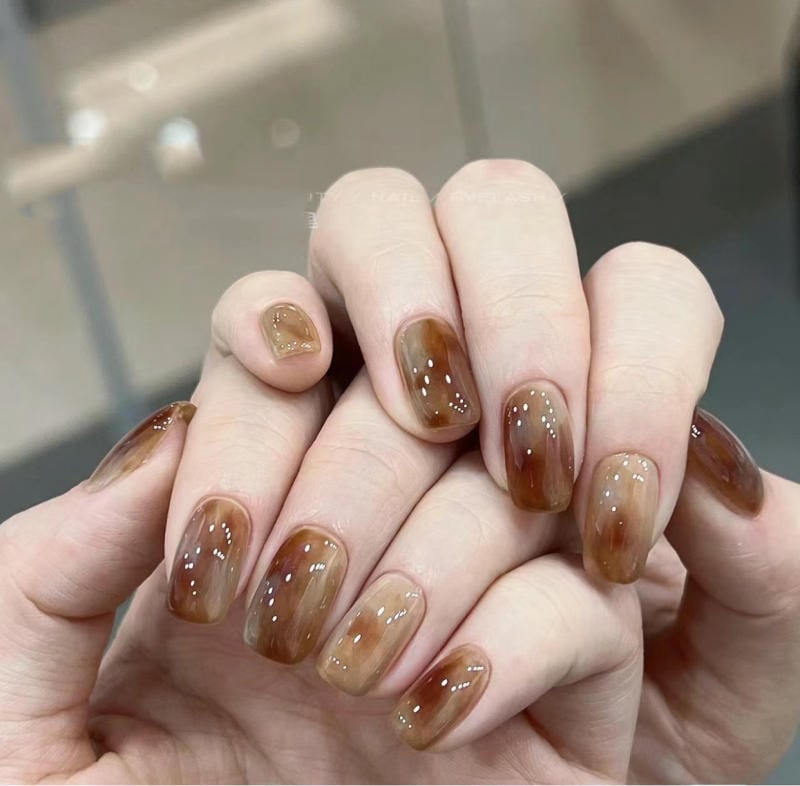 Bàn tay với móng tay được sơn màu nâu bóng, phản chiếu ánh sáng, trên nền không gian mờ.