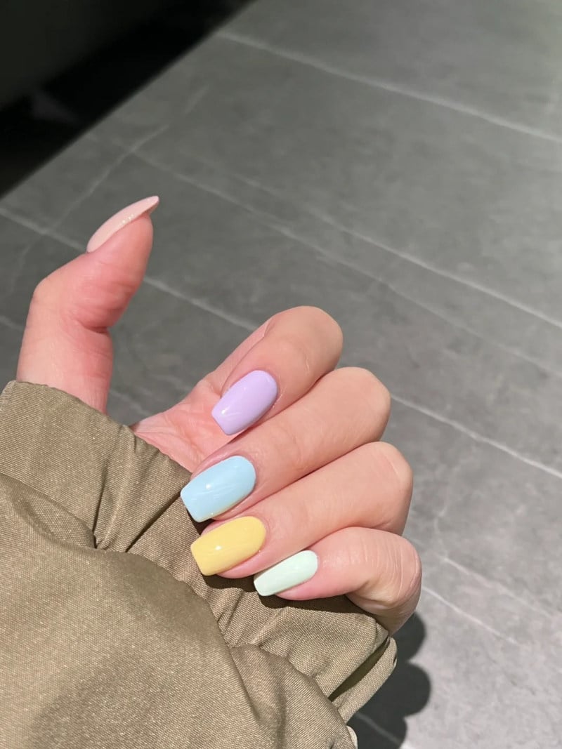 Tay với móng tay sơn màu pastel gồm tím, xanh, vàng, xanh nhạt, trên nền gạch màu xám.