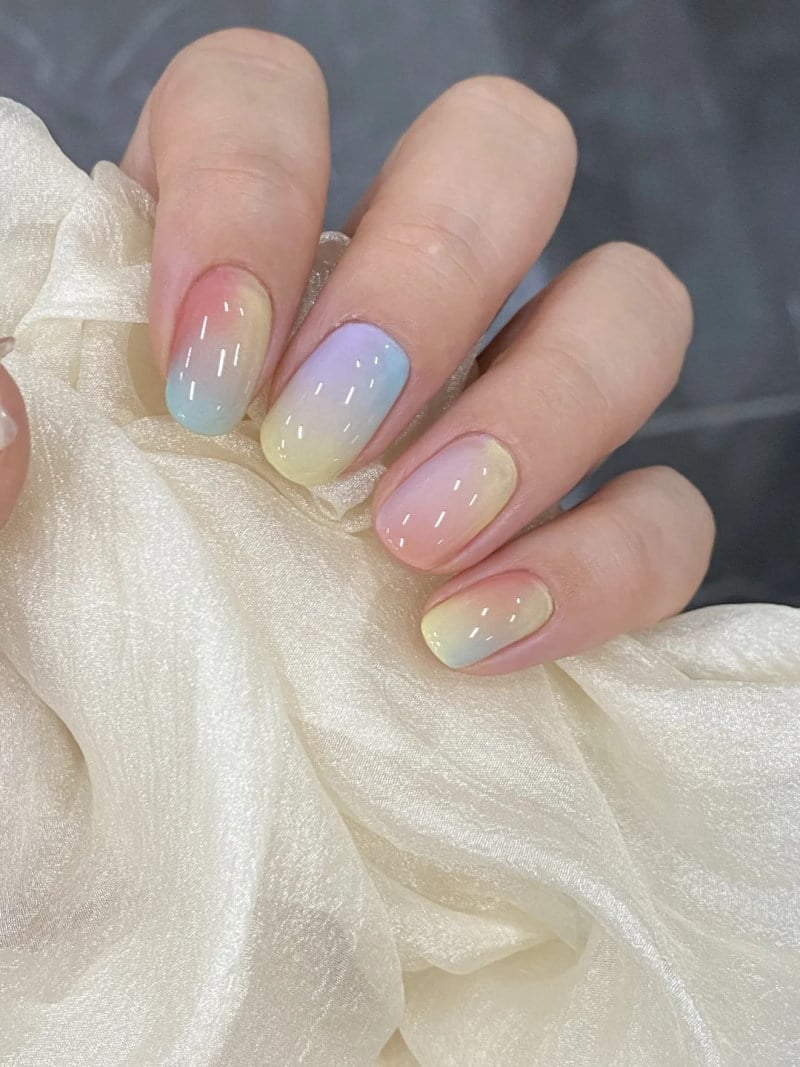 Bàn tay với móng tay sơn ombre màu pastel, cầm một mảnh vải sáng màu, nền tối.