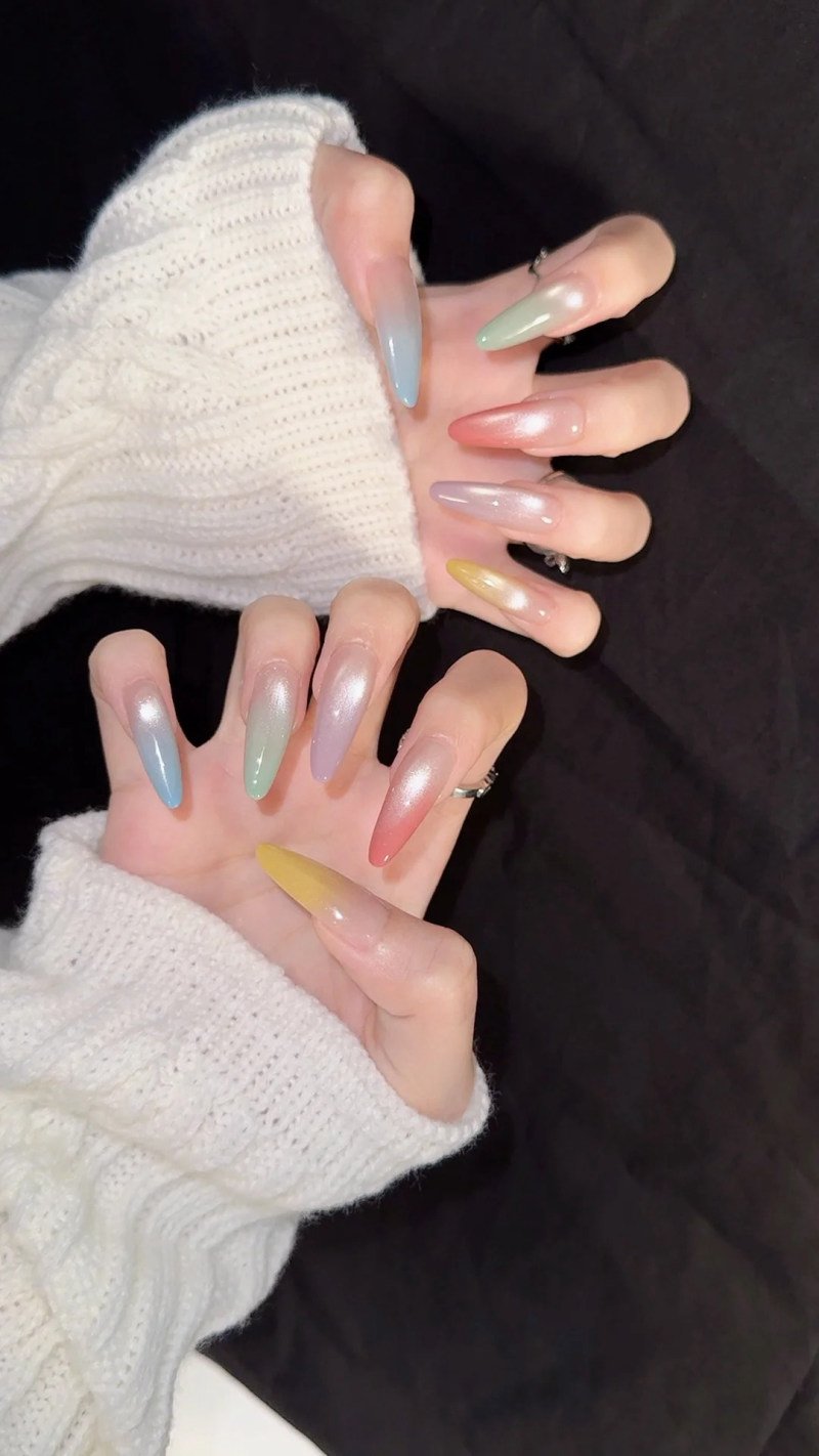Hai bàn tay với móng tay dài, sơn màu pastel khác nhau, áo len trắng, nền tối.