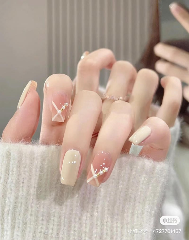 Bàn tay với móng tay được sơn màu be và trang trí họa tiết đính đá lấp lánh, nền áo len màu trắng.