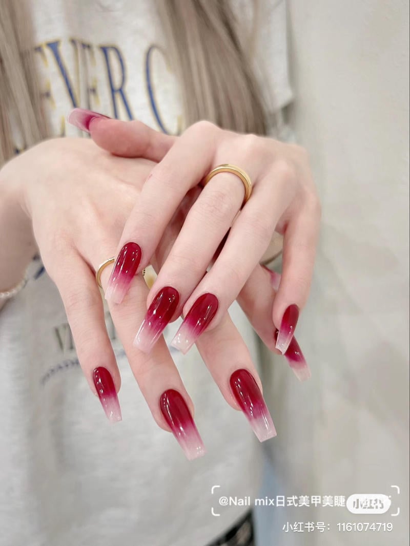 Bàn tay với móng tay dài sơn màu đỏ ombre, đeo nhẫn vàng.