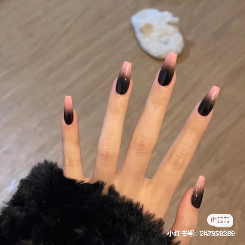 Bàn tay với móng tay sơn màu đen và hồng ombre, nền là sàn gỗ và một vật thể màu trắng.