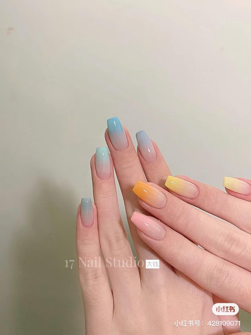 Bàn tay với móng tay sơn màu ombre xanh và vàng cam, trên nền tường màu sáng.