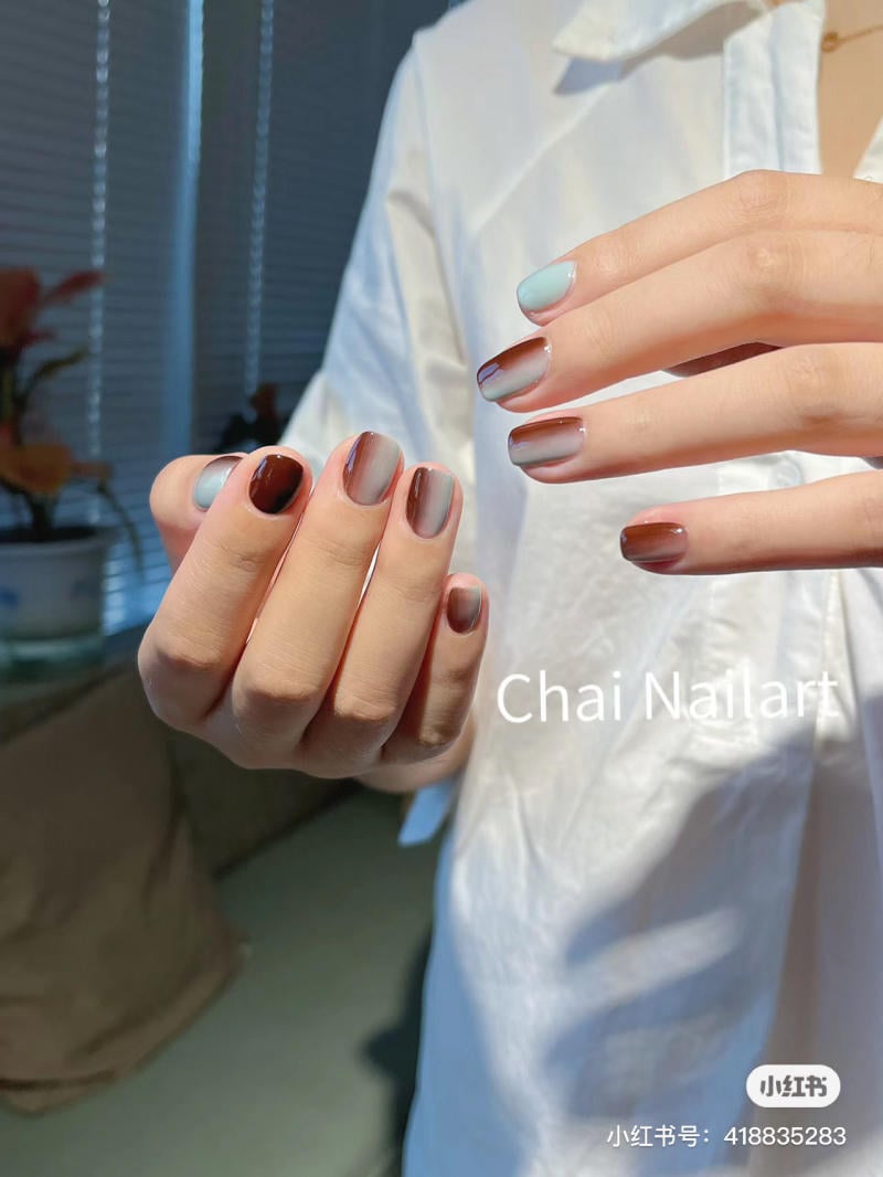Bàn tay với móng tay được sơn màu nâu và xanh nhạt, mặc áo sơ mi trắng, nền mờ.