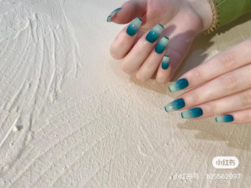 Bàn tay với móng tay sơn màu xanh ombre, nền bề mặt trắng có kết cấu.
