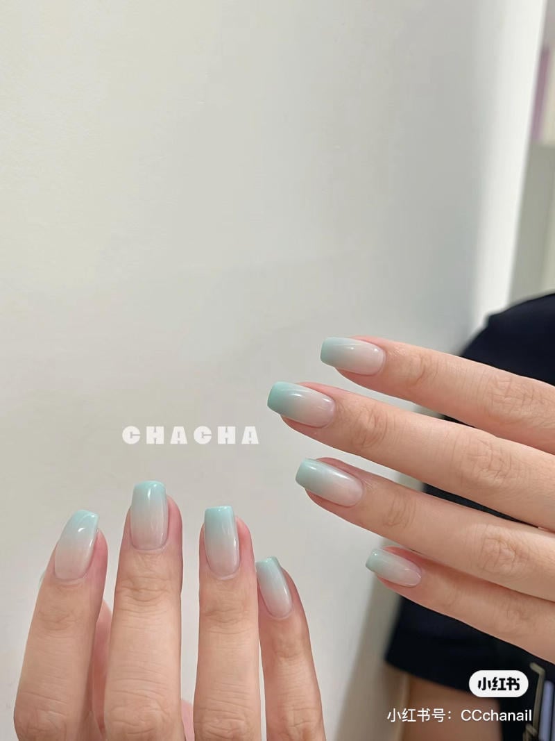 Bàn tay với móng tay được sơn màu xanh nhạt ombre, nền tường trắng.
