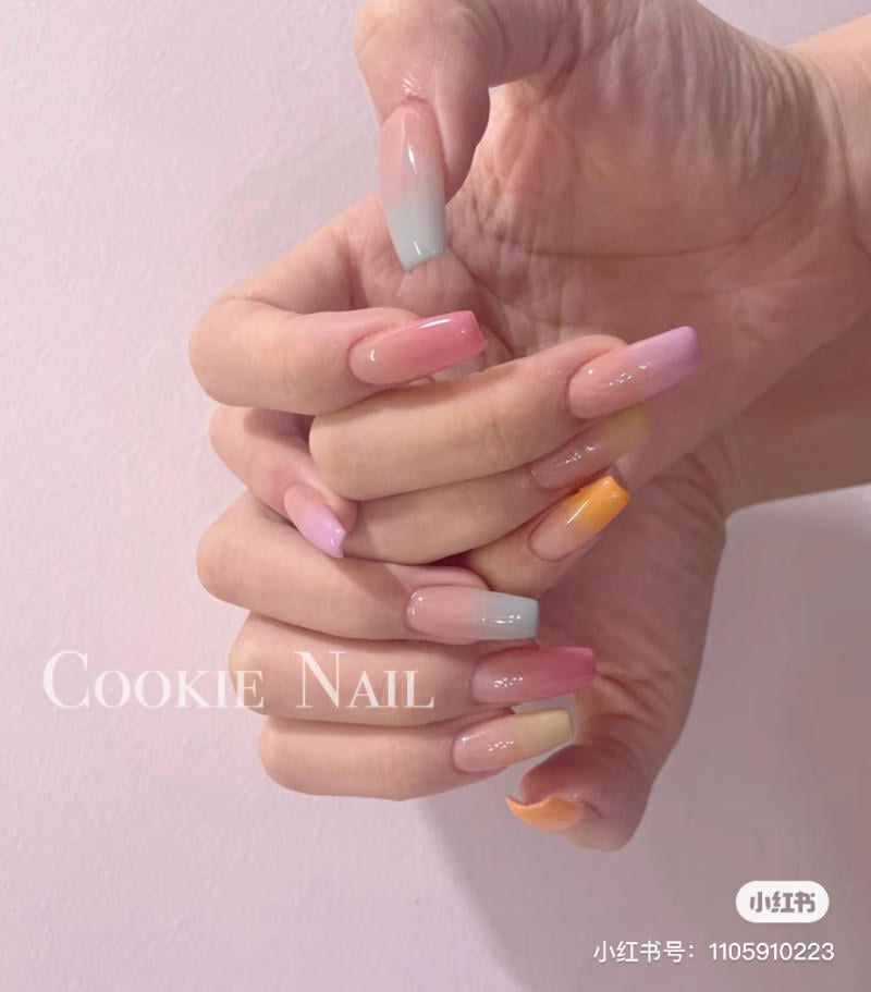 Bàn tay với móng tay dài, được sơn màu ombre nhiều sắc thái hồng, cam và xanh nhạt, trên nền tường màu hồng.