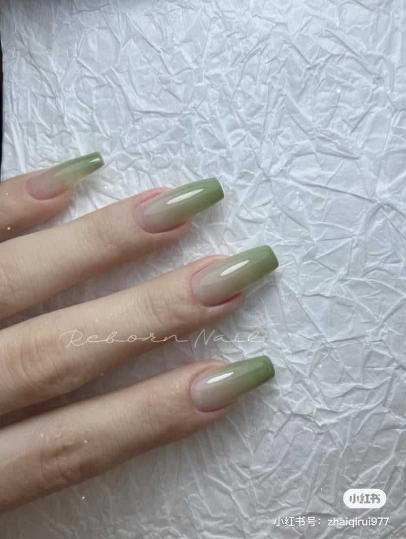 Bàn tay với móng tay sơn màu xanh lá ombre trên nền giấy trắng có kết cấu nhăn.