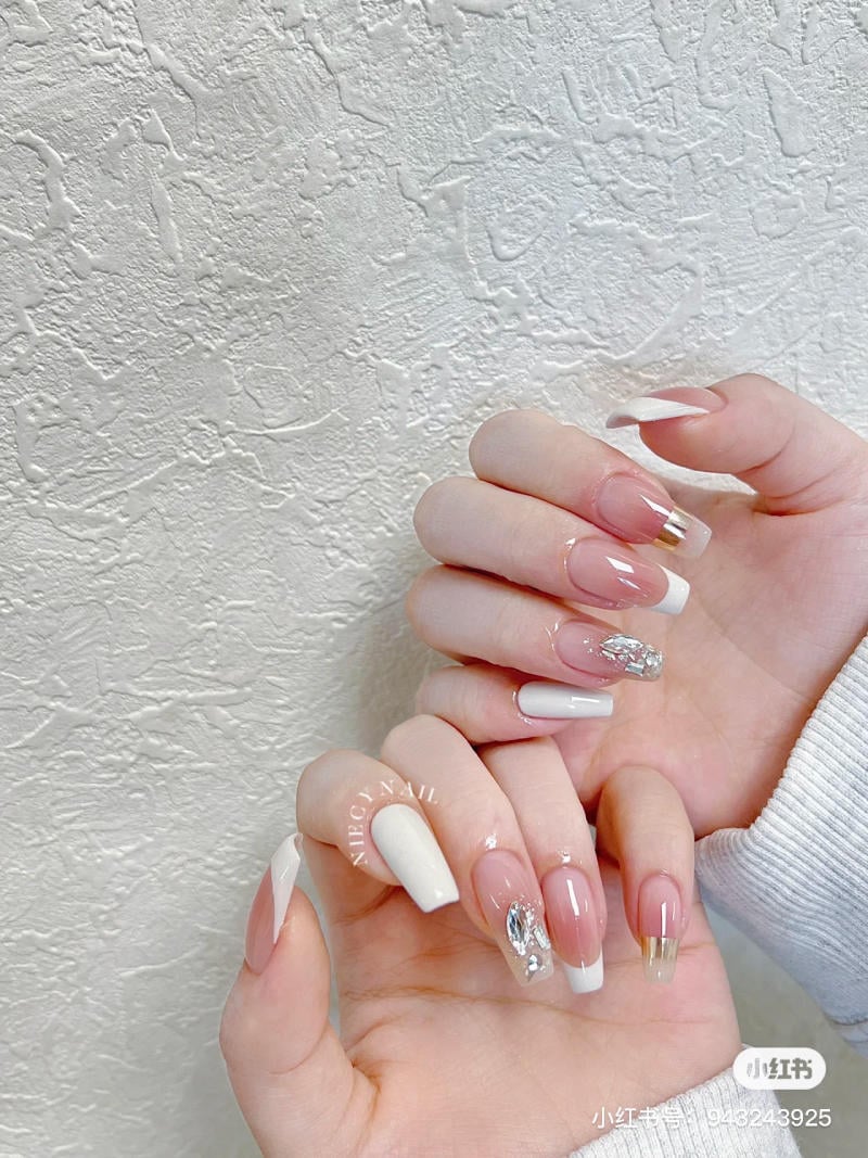Bàn tay với móng tay được trang trí nghệ thuật, có màu trắng và viền bạc, trên nền tường màu trắng.