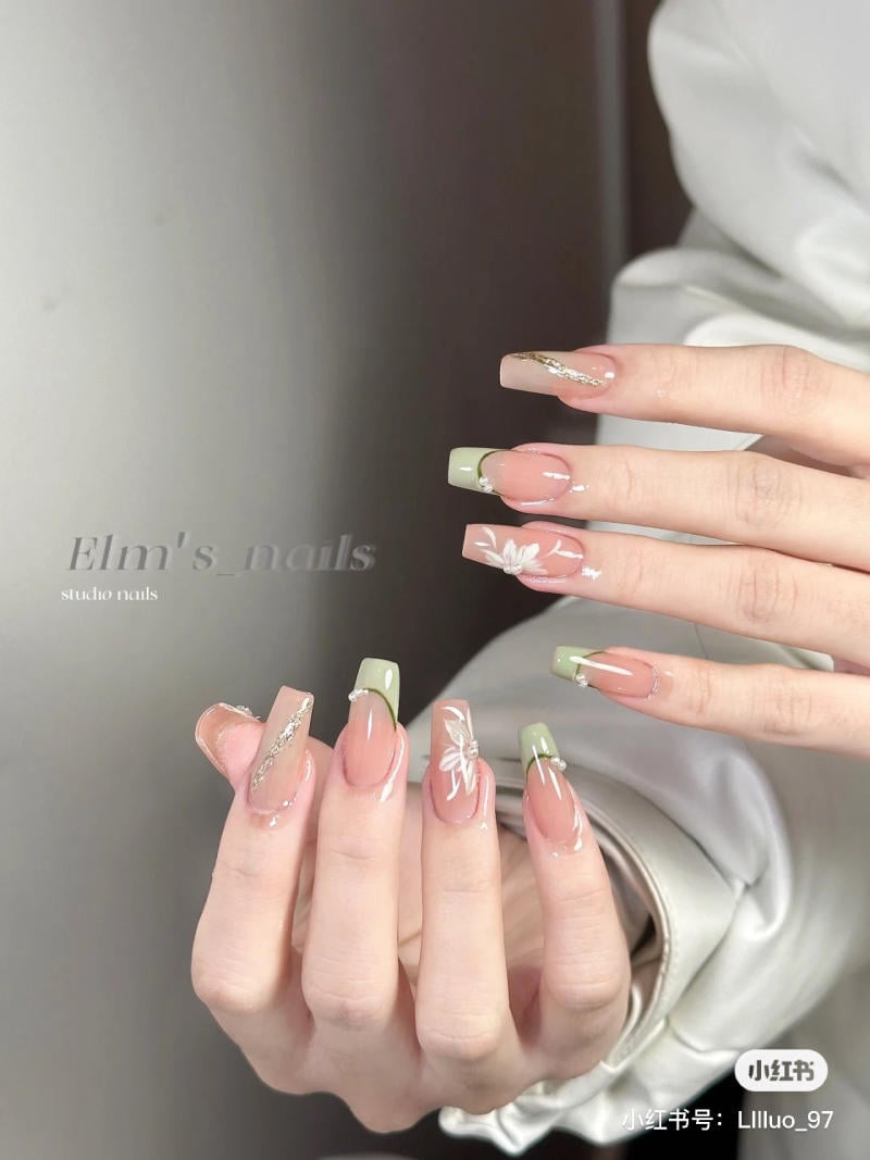 Bàn tay với móng tay dài được sơn màu hồng nhạt và xanh lá, trang trí hoa và họa tiết lấp lánh.