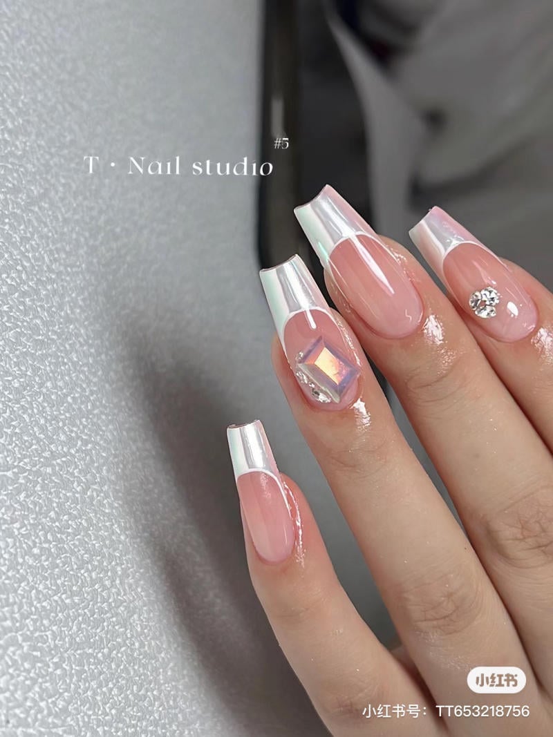 Bàn tay với móng tay được trang trí họa tiết ánh kim và đính đá lấp lánh, nền màu hồng nhẹ nhàng.