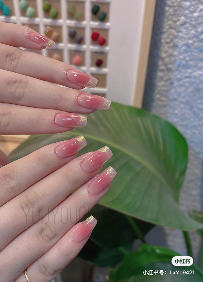 Bàn tay với móng tay được sơn màu hồng nhạt, phía sau là bảng màu và lá cây.