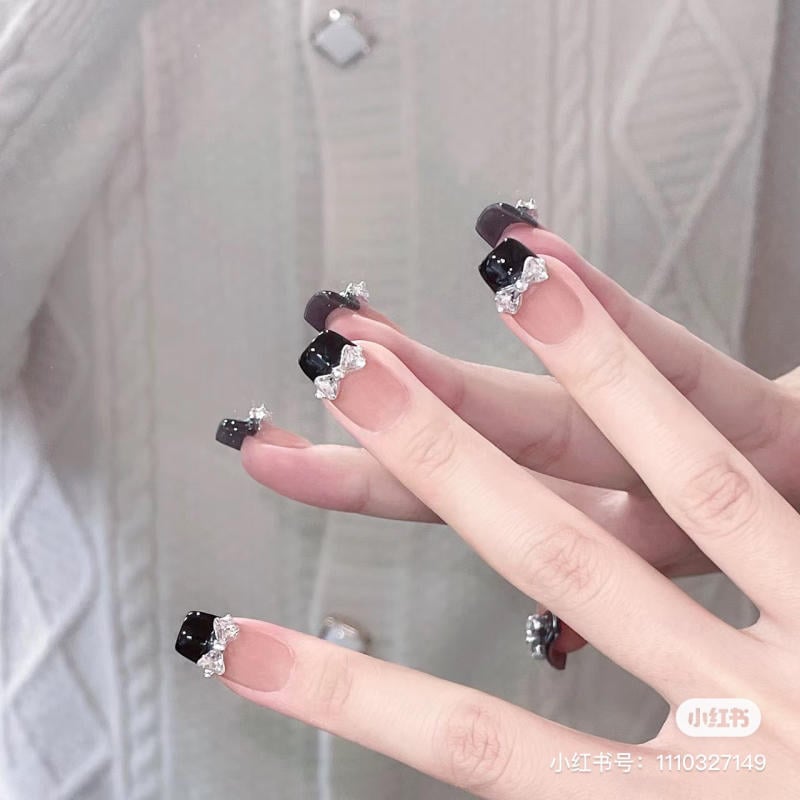 Bàn tay với móng tay đen trang trí nơ bạc, nền áo len trắng.