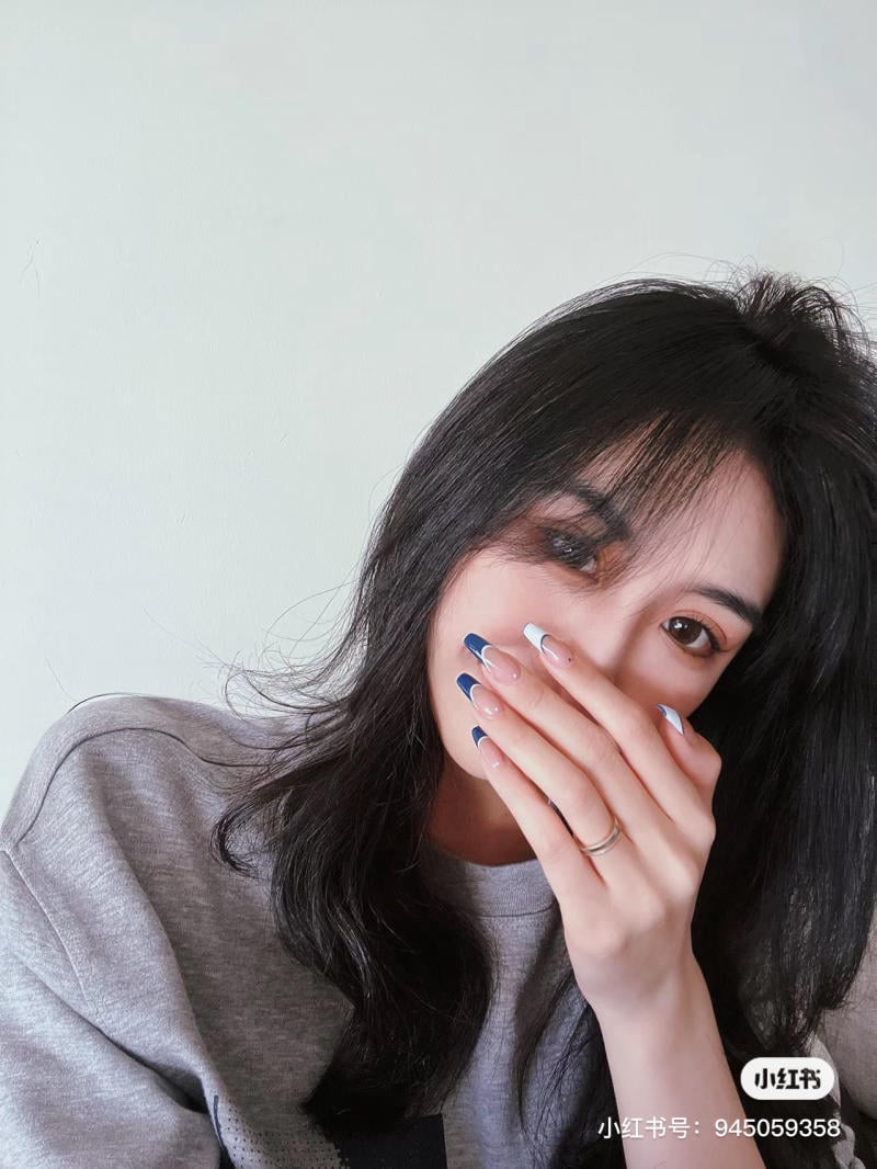 Người phụ nữ tóc đen mặc áo xám, che miệng bằng tay có móng sơn màu xanh và trắng.