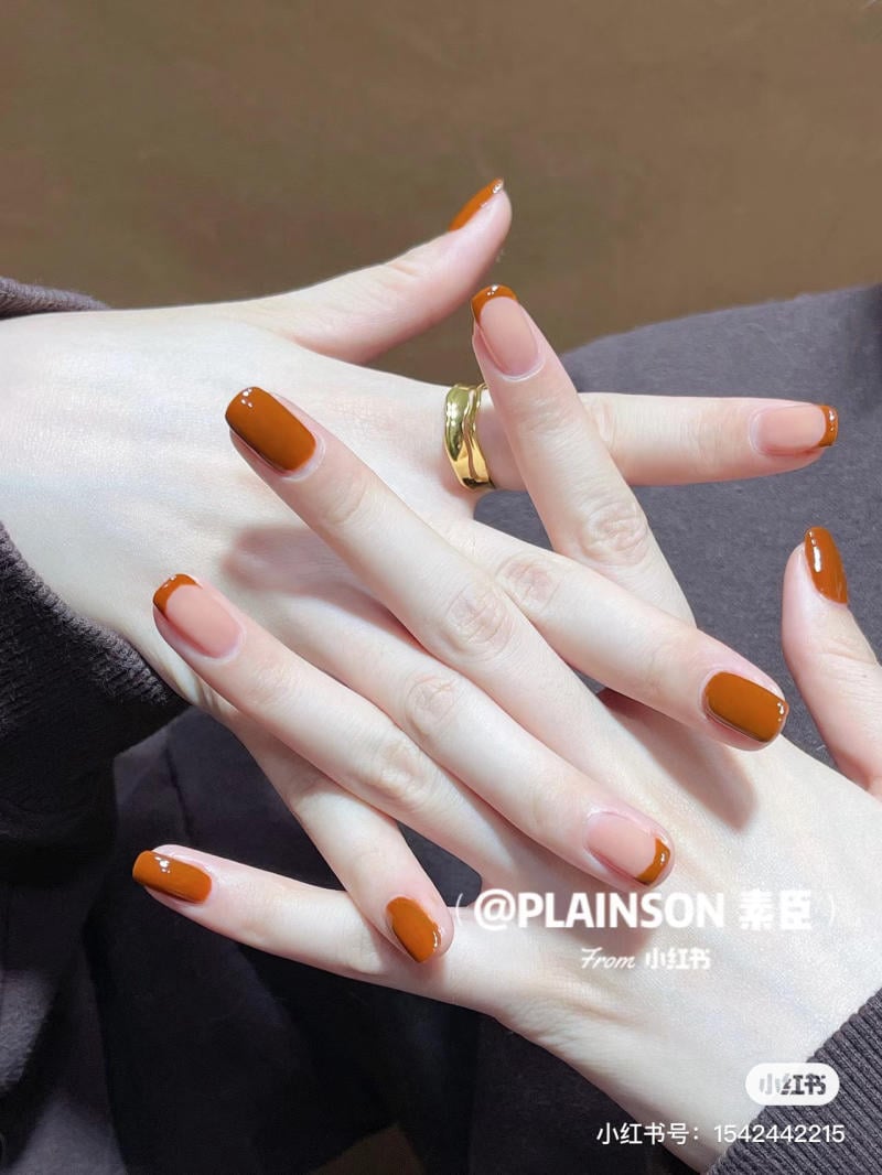Bàn tay với móng tay sơn màu nâu cam, đeo nhẫn vàng, trên nền vải màu tối.