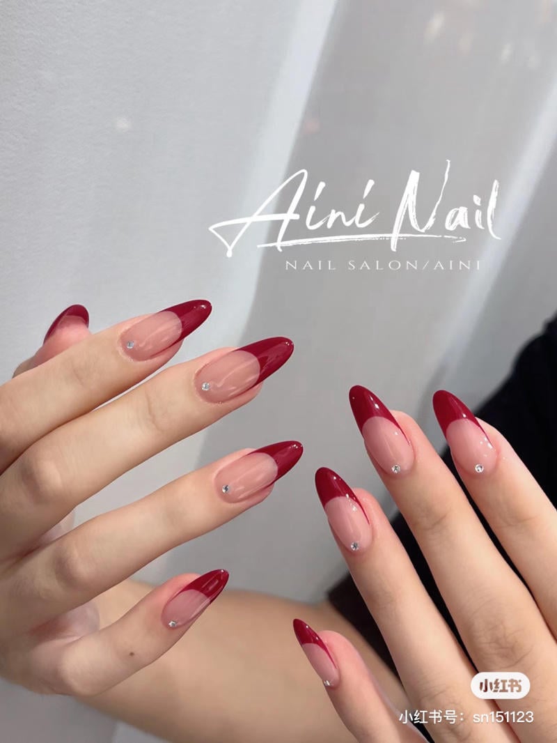 Bàn tay với móng tay sơn màu đỏ và đính đá nhỏ, nền trắng, chữ "Aini Nail" trên hình.
