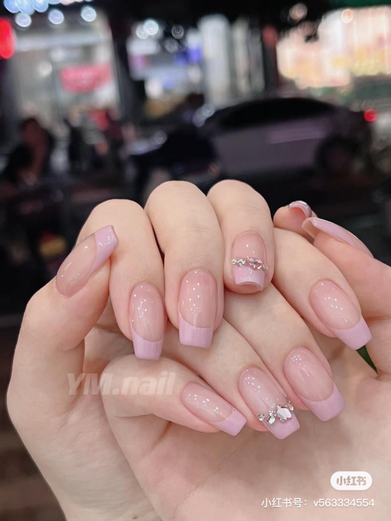 Bàn tay với móng tay được sơn màu hồng nhạt, một số móng trang trí đá lấp lánh, nền mờ nhạt phía sau.