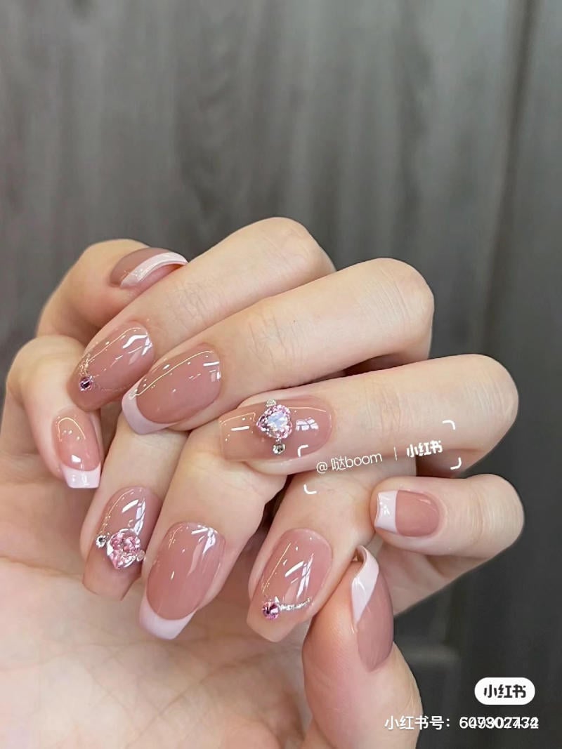 Bàn tay với móng tay được sơn màu hồng nude, trang trí đá và viền trắng, trên nền xám nhạt.