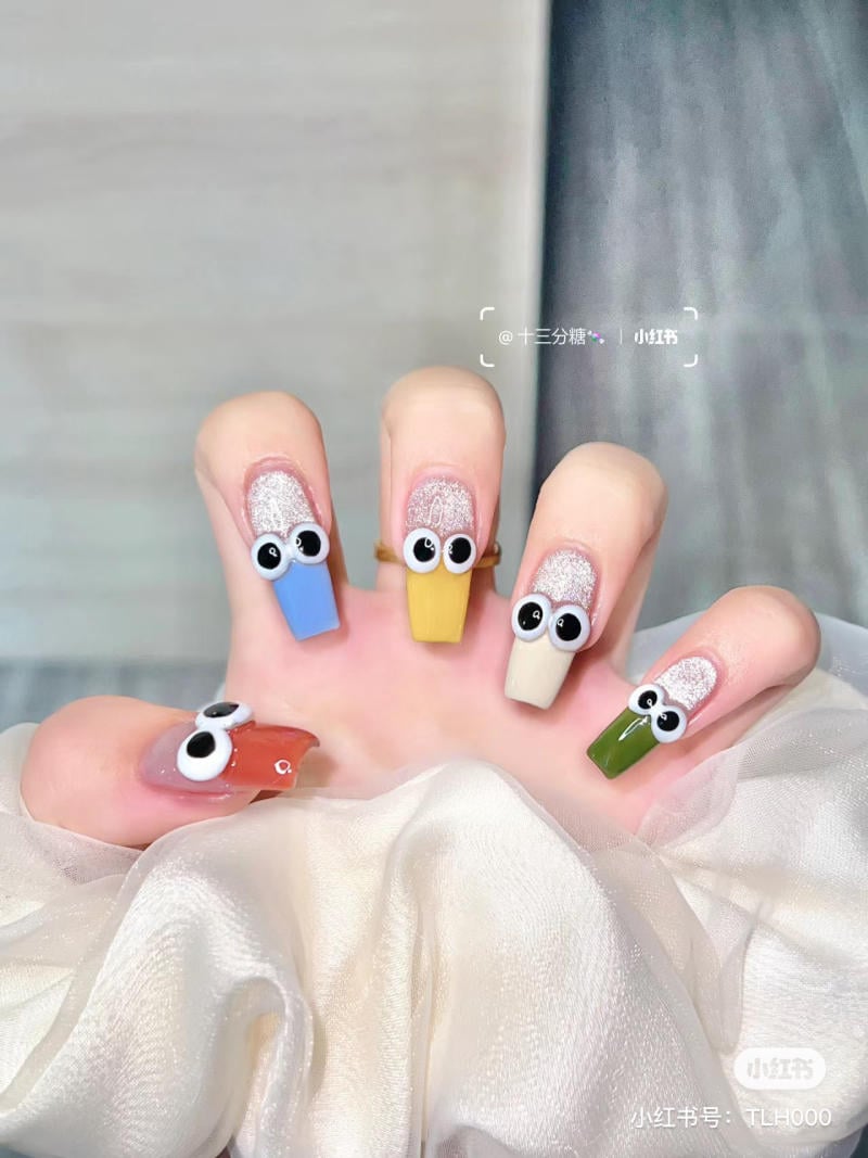 Bàn tay với móng tay sơn màu sắc khác nhau, có hình mắt giả trang trí, đặt trên nền vải trắng.