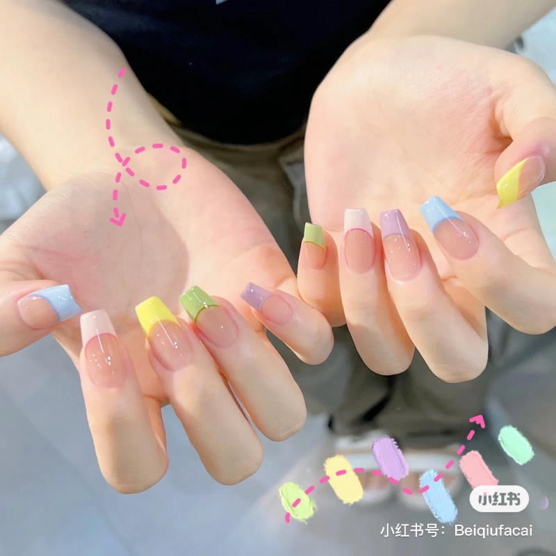 Bàn tay với móng tay dài, được sơn đầu móng nhiều màu sắc tươi sáng, bao gồm xanh, vàng, tím, và hồng.