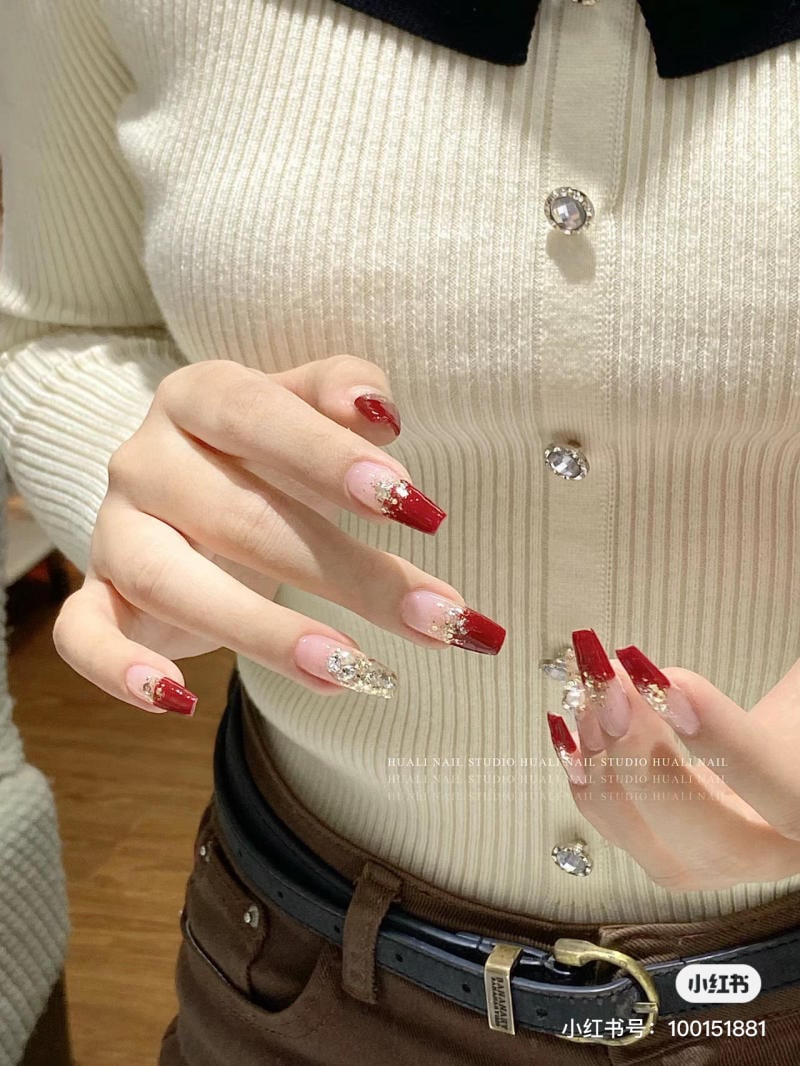 Người mặc áo trắng, tay có móng đỏ và vàng lấp lánh, đứng trước nền gỗ.