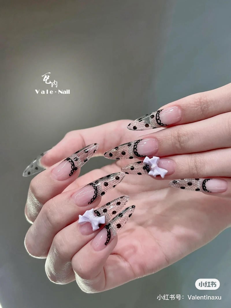 Bàn tay với móng tay dài, trang trí hoa văn ren đen và nơ trắng nổi bật trên nền màu nude.