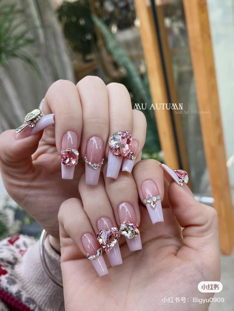 Bàn tay với móng tay giả dài, trang trí đá quý màu hồng và bạc, nền cảnh thiên nhiên mờ nhạt phía sau.