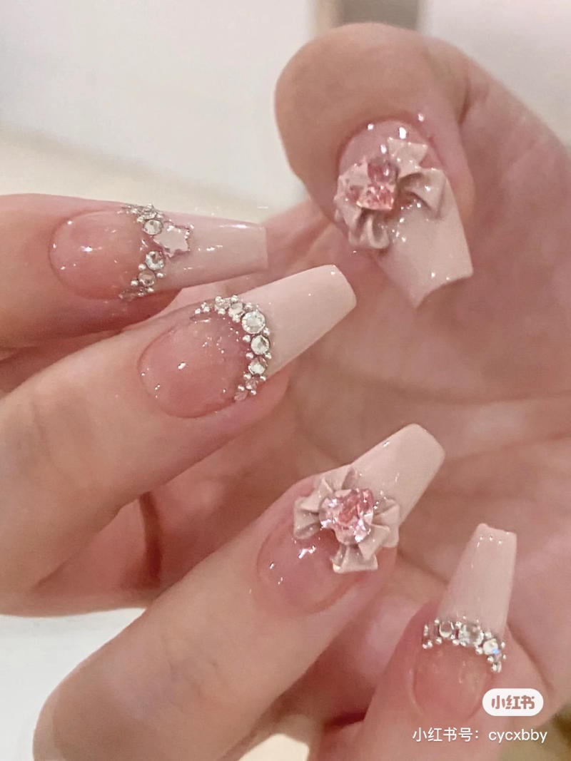 Bàn tay với móng tay dài, sơn màu hồng nhạt, trang trí đá và hoa nổi bật.